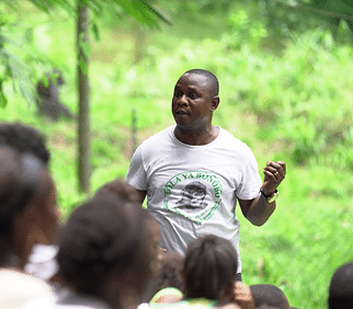 Man in a white t-shirt speaks to a group outdoors, surrounded by lush greenery. T-shirt has "LOLA YA BONOBO" and an image printed on it.