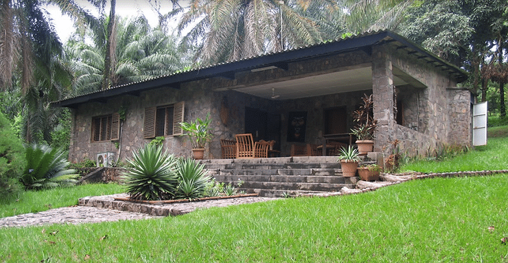 Stone house with porch, surrounded by lush greenery and palm trees. Wooden chairs and potted plants on the steps. Peaceful, tropical setting.