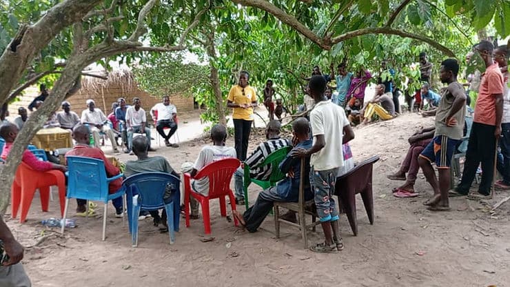 Community meeting under trees with black African people seated in colorful chairs. A person in a yellow shirt standing in center of circle speaks.