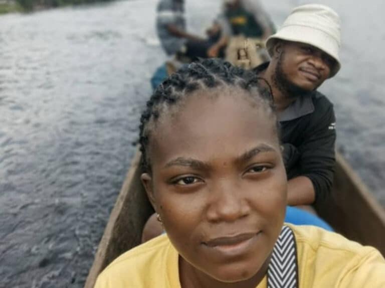 Two people on a long canoe in a river. The woman in front, wearing yellow, is taking a selfie. The man behind wears a bucket hat.