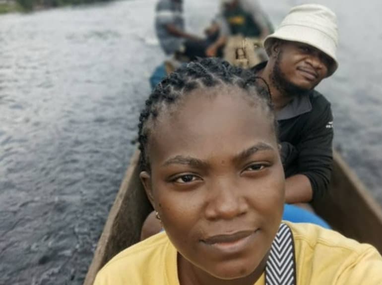 Two people on a long canoe in a river. The woman in front, wearing yellow, is taking a selfie. The man behind wears a bucket hat.