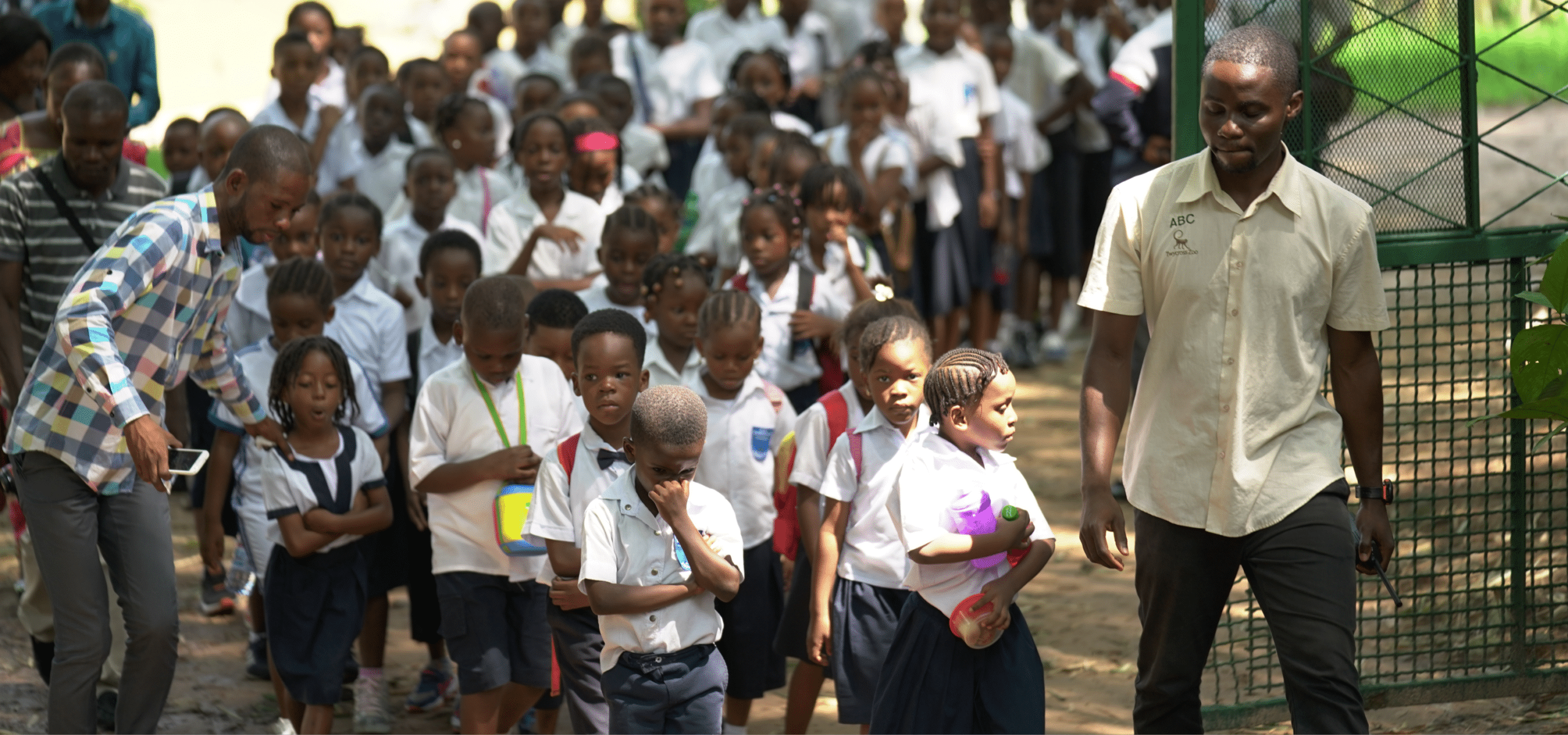 A large group of school children wearing white shirts walks with two adult male teachers during a conservation education program at Lola ya Bonobo sanctuary.